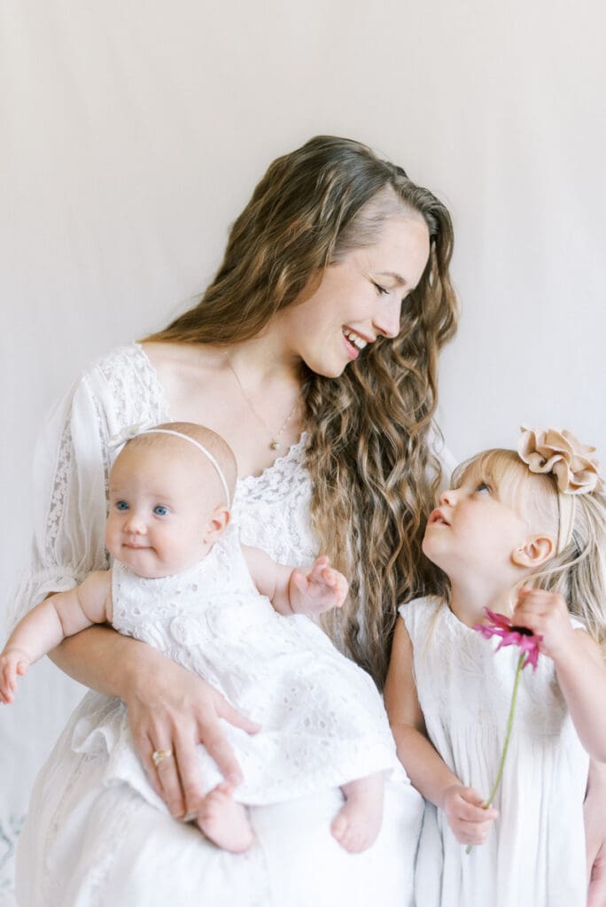 A mother dressed in white looks lovingly at her two daughters in a studio portrait photo session